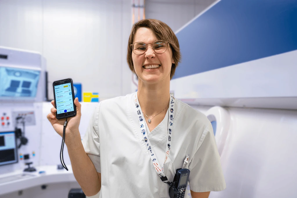 Smiling nurse in scrubs holding a smartphone running PMcardio in a hospital